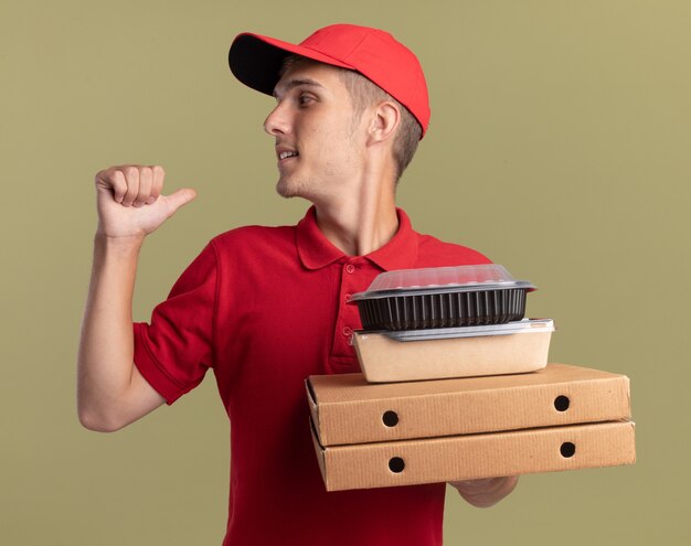 Heureux jeune livreur blonde détient des emballages alimentaires sur des boîtes de pizza et des points en arrière sur vert olive