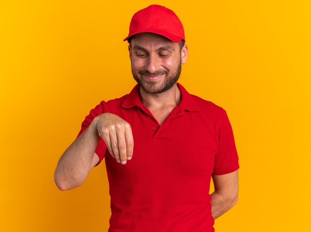 Heureux jeune livreur caucasien en uniforme rouge et casquette gardant la main derrière le dos faire semblant de tenir quelque chose en regardant sa main isolée sur un mur orange