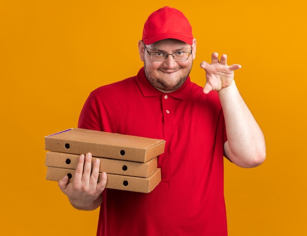 Heureux jeune livreur en surpoids dans des lunettes optiques tenant des boîtes à pizza et gesticulant une patte de tigre isolée sur un mur orange avec espace pour copie