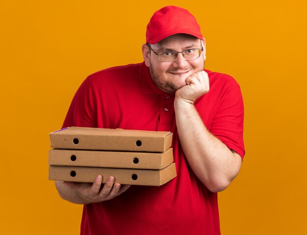 Heureux jeune livreur en surpoids dans des lunettes optiques tenant des boîtes à pizza et mettant la main sur le menton isolé sur un mur orange avec espace de copie