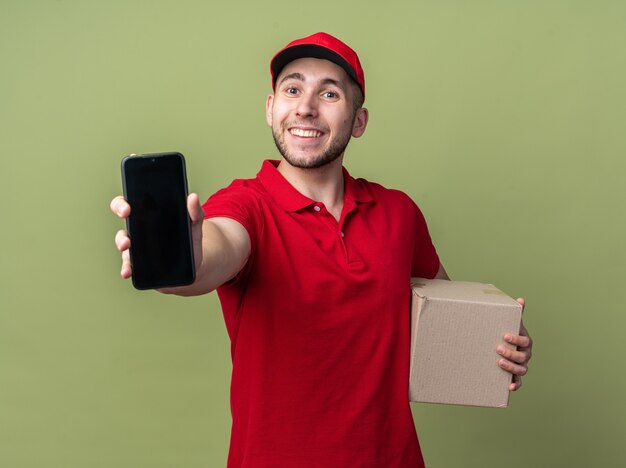 Heureux jeune livreur en uniforme, tenant une boîte en carton et montrant un smartphone