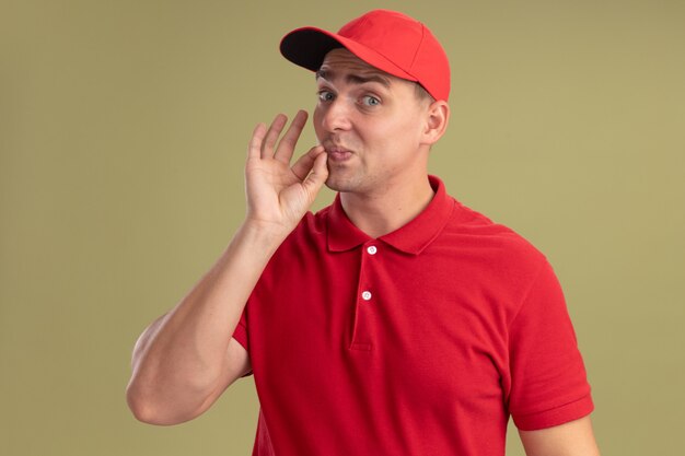 Heureux jeune livreur vêtu d'un uniforme et d'une casquette montrant un geste délicieux isolé sur un mur vert olive