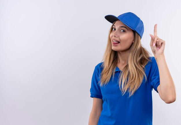 Heureux jeune livreuse portant l'uniforme bleu et une casquette montrant la langue et pointe vers le haut isolé sur un mur blanc avec copie espace