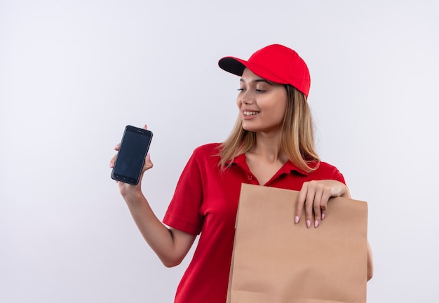 Heureux jeune livreuse portant l'uniforme rouge et une casquette tenant un sac en papier et regardant le téléphone dans sa main isolé sur fond blanc