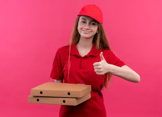 Heureux jeune livreuse en uniforme rouge tenant des paquets et montrant le pouce vers le haut sur l'espace rose isolé