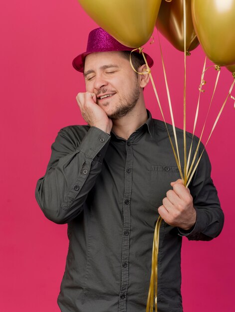 Heureux jeune mec aux yeux fermés portant un chapeau rose tenant des ballons mettant la main sur la joue isolé sur rose