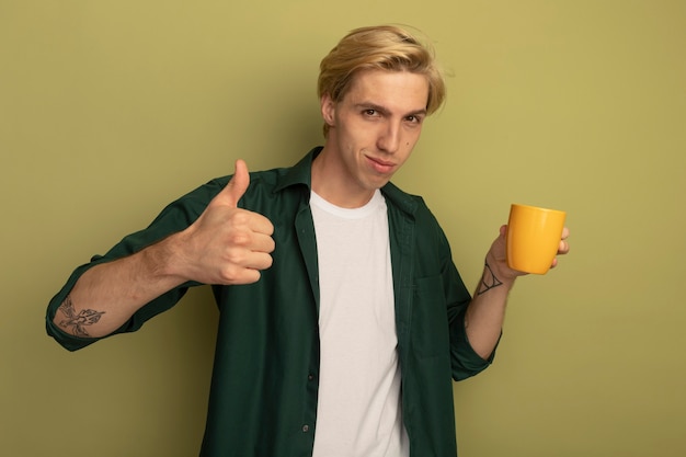 Heureux jeune mec blond portant un t-shirt vert tenant une tasse de thé et montrant le pouce vers le haut