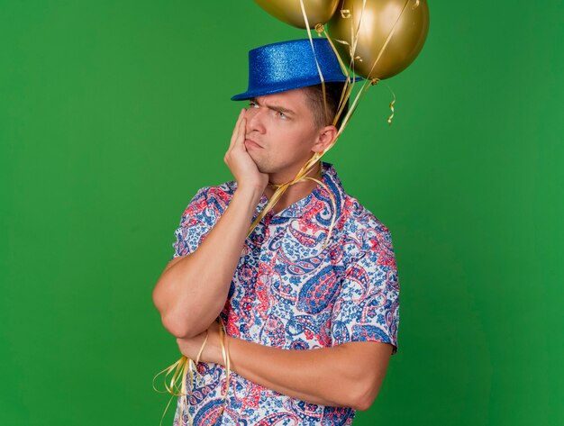 Heureux jeune mec de la fête à côté portant un chapeau bleu tenant des ballons attachés autour du cou mettant la main sous le menton isolé sur vert