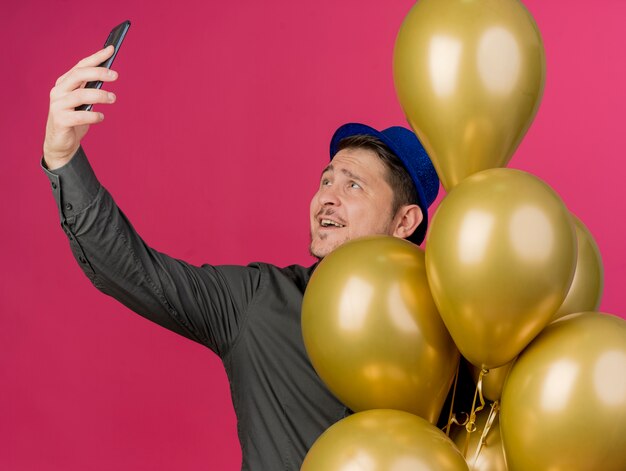 Heureux jeune mec portant chapeau bleu debout parmi les ballons et prendre un selfie isolé sur rose