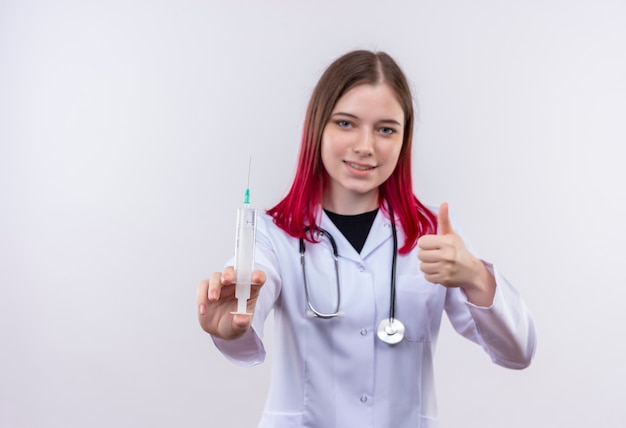 Heureux jeune médecin fille portant robe médicale stéthoscope tenant la seringue son pouce vers le haut sur fond blanc isolé