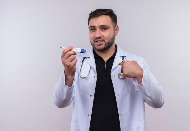 Heureux jeune médecin de sexe masculin barbu portant blouse blanche avec stéthoscope tenant un thermomètre numérique souriant confiant pointant vers lui-même