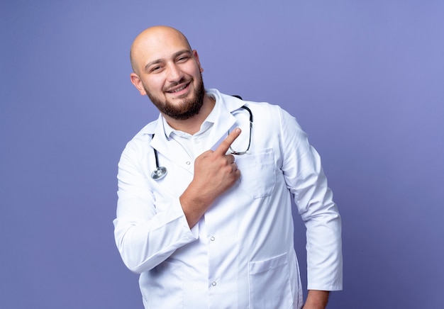 Heureux jeune médecin de sexe masculin chauve portant une robe médicale et des points de stéthoscope sur le côté isolé sur fond bleu avec espace de copie