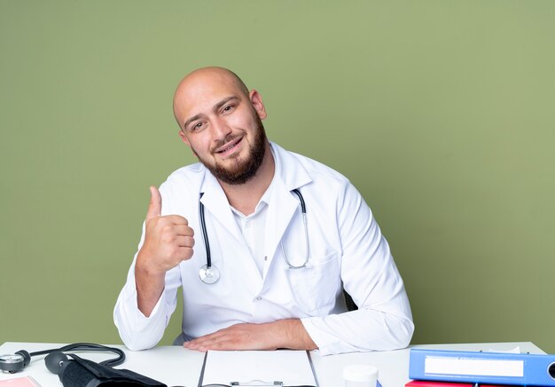 Heureux jeune médecin de sexe masculin chauve portant une robe médicale et un stéthoscope assis au bureau de travail avec des outils médicaux son pouce vers le haut isolé sur fond vert