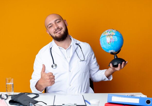 Heureux jeune médecin de sexe masculin chauve portant une robe médicale et un stéthoscope assis au bureau de travail avec des outils médicaux tenant globe son pouce vers le haut isolé sur fond orange