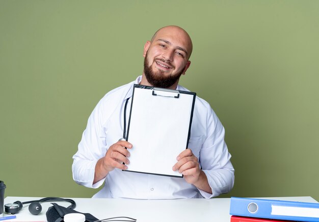 Photo gratuite heureux jeune médecin de sexe masculin chauve portant une robe médicale et un stéthoscope assis au bureau de travail avec des outils médicaux tenant le presse-papiers isolé sur fond vert