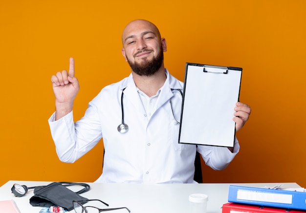 Photo gratuite heureux jeune médecin de sexe masculin chauve portant une robe médicale et un stéthoscope assis au bureau de travail avec des outils médicaux tenant le presse-papiers et les points en haut isolé sur fond orange