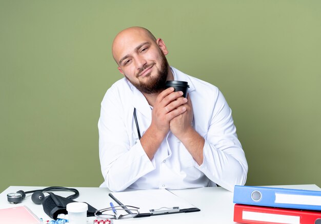 Heureux jeune médecin de sexe masculin chauve portant robe médicale et stéthoscope assis au travail de bureau avec des outils médicaux tenant une tasse de café isolé sur fond vert