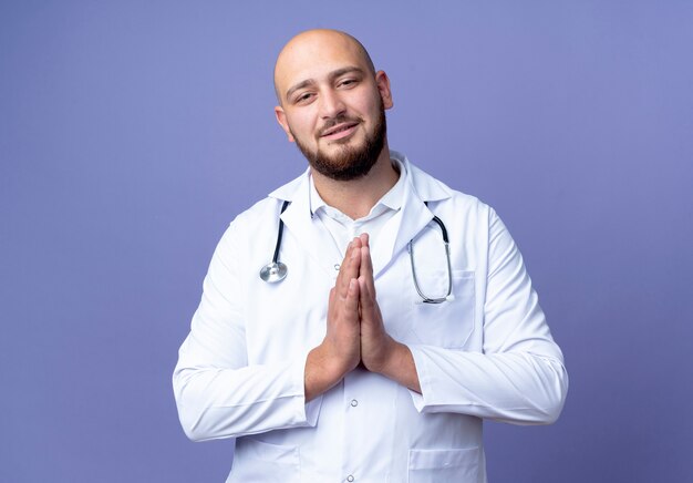 Heureux jeune médecin de sexe masculin chauve portant une robe médicale et un stéthoscope montrant le geste de prier isolé sur fond bleu