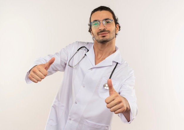 Heureux jeune médecin de sexe masculin avec des lunettes optiques portant une robe blanche avec stéthoscope ses pouces vers le haut sur un mur blanc isolé avec copie espace