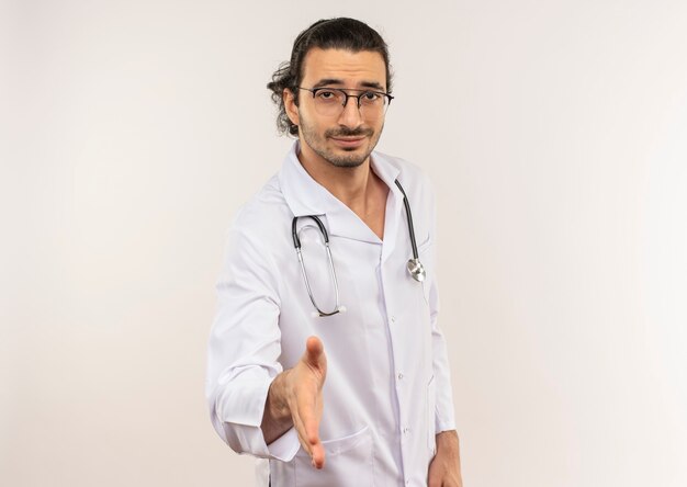 Heureux jeune médecin de sexe masculin avec des lunettes optiques portant une robe blanche avec stéthoscope tenant la main sur un mur blanc isolé avec copie espace