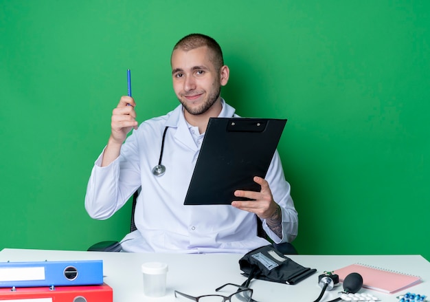 Heureux jeune médecin de sexe masculin portant une robe médicale et un stéthoscope assis au bureau avec des outils de travail tenant le presse-papiers et un stylo isolé sur un mur vert