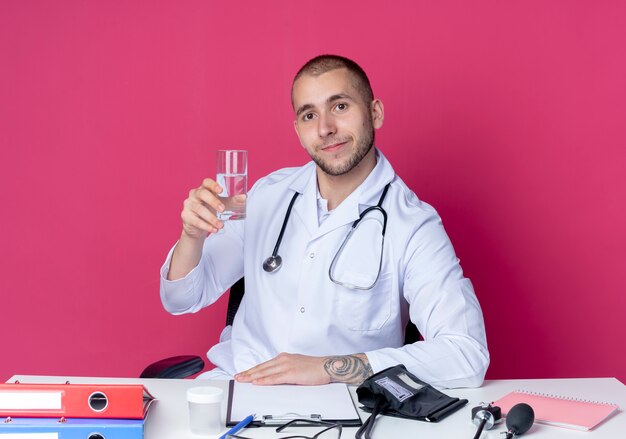 Heureux jeune médecin de sexe masculin portant une robe médicale et un stéthoscope assis au bureau avec des outils de travail tenant un verre d'eau mettant la main sur un bureau isolé sur un mur rose