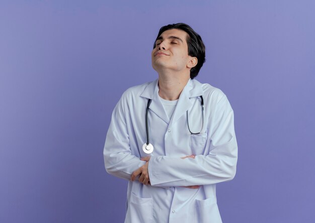 Heureux jeune médecin de sexe masculin portant une robe médicale et un stéthoscope en gardant la main sur le ventre et une autre main sur le bras avec les yeux fermés isolé sur un mur violet avec espace de copie