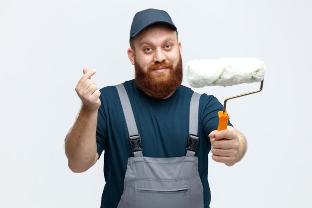 Heureux jeune ouvrier du bâtiment portant une casquette et un uniforme regardant la caméra étirant le rouleau de peinture vers la caméra montrant un geste d'argent isolé sur fond blanc