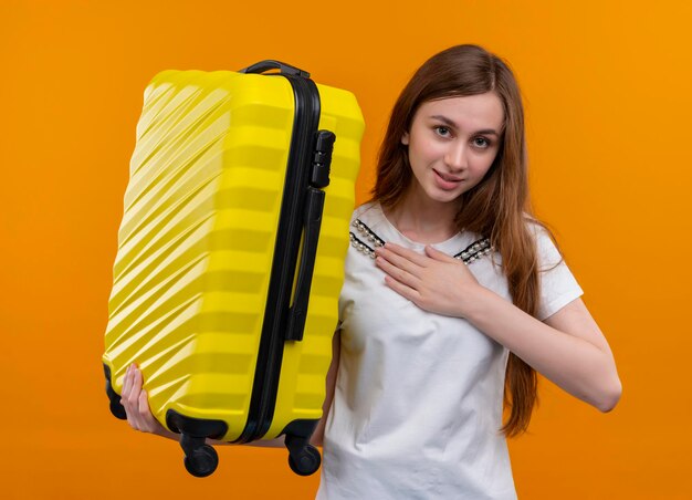 Heureux jeune voyageur fille tenant valise et mettant la main sur la poitrine sur l'espace orange isolé