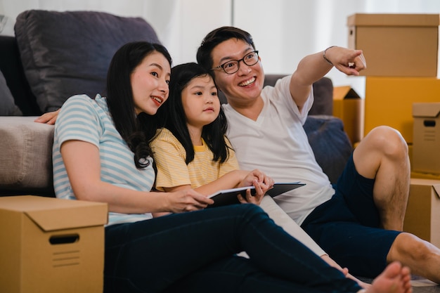 Heureux jeunes propriétaires asiatiques ont acheté une nouvelle maison. Maman chinoise, papa et sa fille s'embrassent dans une nouvelle maison après avoir déménagé, assis sur le sol avec des boîtes ensemble.