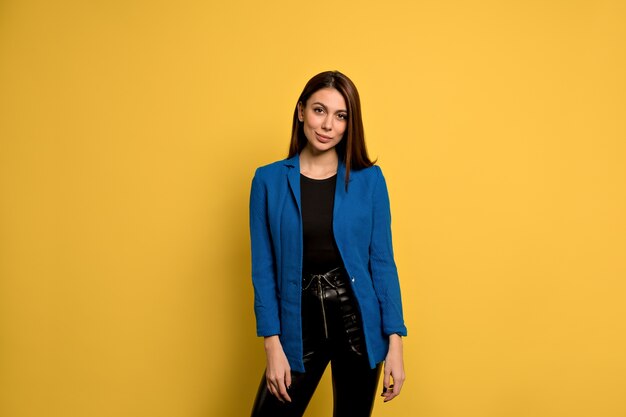 Heureux jolie femme aux cheveux longs, yeux foncés et peau saine vêtue d'une veste bleue, souriant tout en posant contre le mur de béton jaune. Les gens et le style de vie