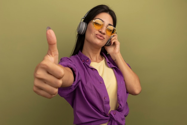 Heureux jolie femme brune à lunettes de soleil avec un casque Thumbs up isolé sur mur vert olive