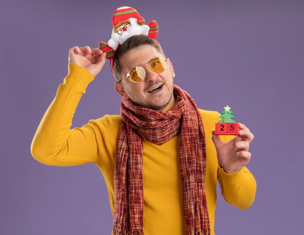 Heureux et joyeux jeune homme en col roulé jaune avec écharpe chaude et lunettes portant une jante drôle avec le père Noël sur la tête montrant des cubes de jouet avec le numéro vingt-cinq debout sur un mur violet
