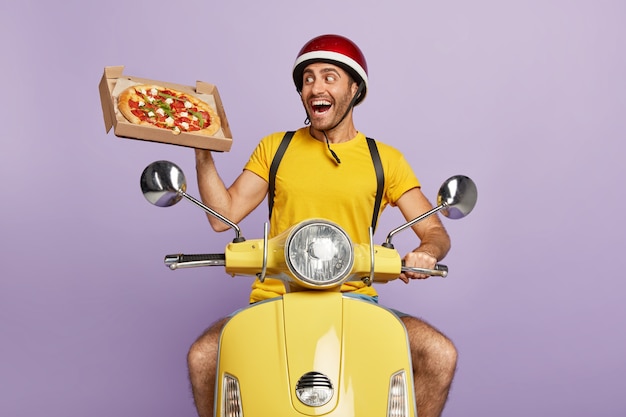 Heureux livreur conduisant un scooter jaune tout en tenant une boîte à pizza