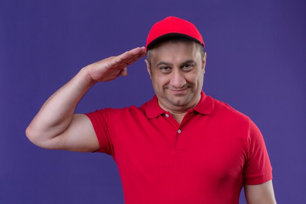 Heureux livreur en uniforme rouge et cap rendant hommage prêt pour le travail souriant sympathique sur mur violet isolé