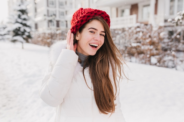 Heureux matin gelé ensoleillé sur l'heure d'hiver de joyeuse jeune femme au chapeau rouge, avec de longs cheveux bruns s'amusant sur la rue pleine de neige.