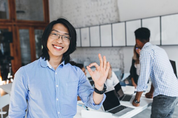 Heureux mec asiatique en t-shirt bleu classique posant avec un signe correct après une conférence avec des collègues. Portrait intérieur d'heureux entrepreneur chinois dans des verres bénéficiant d'une bonne journée.