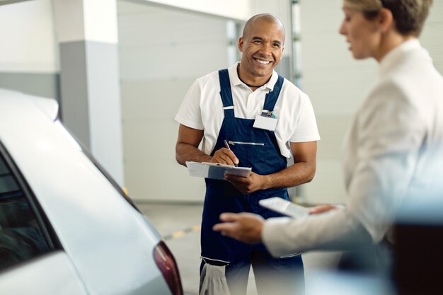 Heureux mécanicien noir prenant des notes tout en parlant à un client dans un atelier de réparation automobile