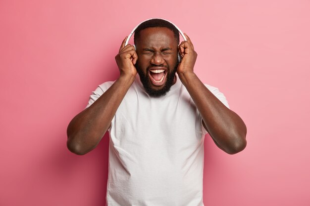 Heureux mélomane écoute la musique préférée dans les écouteurs, bénéficie d'un bon son garde la bouche ouverte et crie fort, porte un t-shirt blanc décontracté, pose en studio contre le mur rose