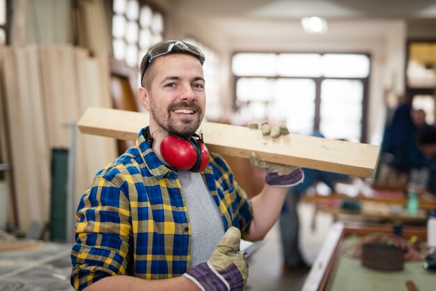 Heureux menuisier souriant tenant du bois et des pouces vers le haut dans son atelier de menuiserie