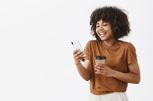 Photo gratuite heureux modèle féminin amical et attrayant à la peau sombre avec des cheveux bouclés tenant une tasse de café en papier et un smartphone en riant tout en lisant une histoire drôle dans un téléphone portable