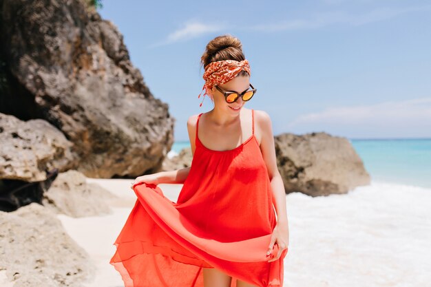 Heureux modèle féminin blanc à lunettes de soleil à la plage avec des rochers. Jolie fille caucasienne jouant avec sa robe rouge près de l'océan.
