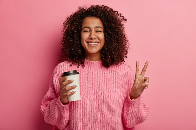 Heureux modèle féminin bouclé à la peau sombre fait un geste de paix, tient un café à emporter, s'amuse pendant la pause, sourit à la caméra, porte un pull, pose sur un mur rose.