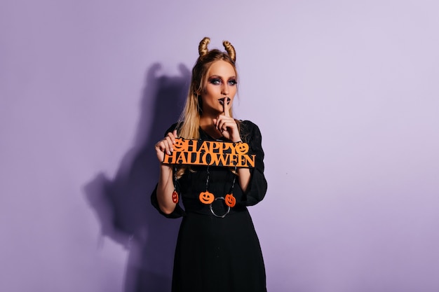 Photo gratuite heureux modèle féminin en élégante robe noire posant à halloween. adorable fille en costume de sorcière debout sur un mur violet.