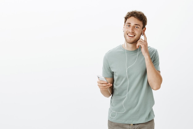 Heureux modèle masculin caucasien avec poils, tenant le smartphone et touchant les écouteurs tout en écoutant de la musique et en profitant du bon son des écouteurs