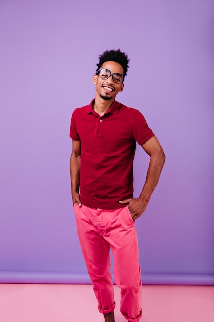 Photo gratuite heureux modèle masculin noir debout dans une pose confiante. homme insouciant souriant en pantalon rose et t-shirt rouge glaçant.