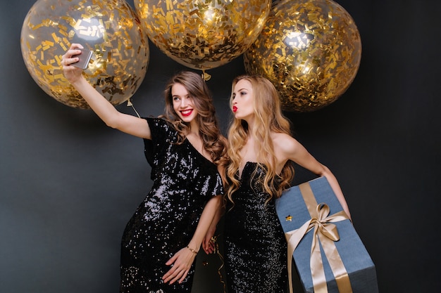 Heureux moments de fête de deux jeunes femmes à la mode faisant selfie. Robe noire de luxe, longs cheveux bouclés, gros ballons avec des guirlandes dorées, présent, s'amusant, souriant.