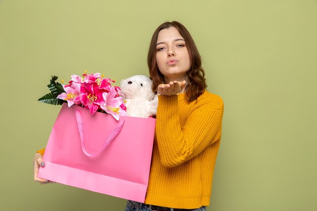 Heureux de montrer le geste de baiser belle jeune fille le jour de la femme heureuse tenant un sac-cadeau isolé sur un mur vert olive