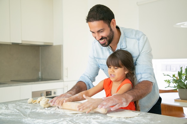 Heureux papa et fille à rouler la pâte sur la table de cuisine avec de la farine en désordre.