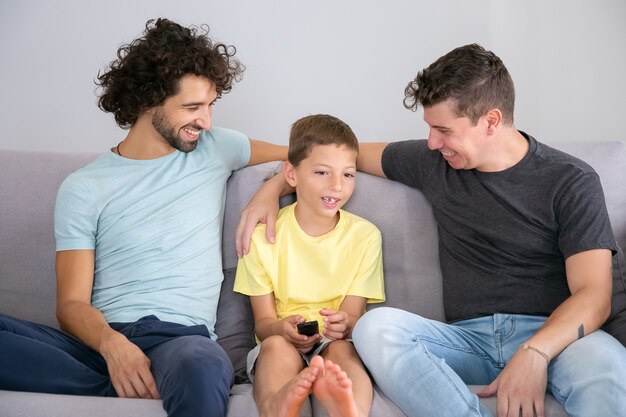Heureux papas et fils gay regardant une émission de télévision discutant ensemble à la maison, assis sur un canapé dans le salon, souriant et étreignant le garçon. Concept de divertissement familial et à domicile